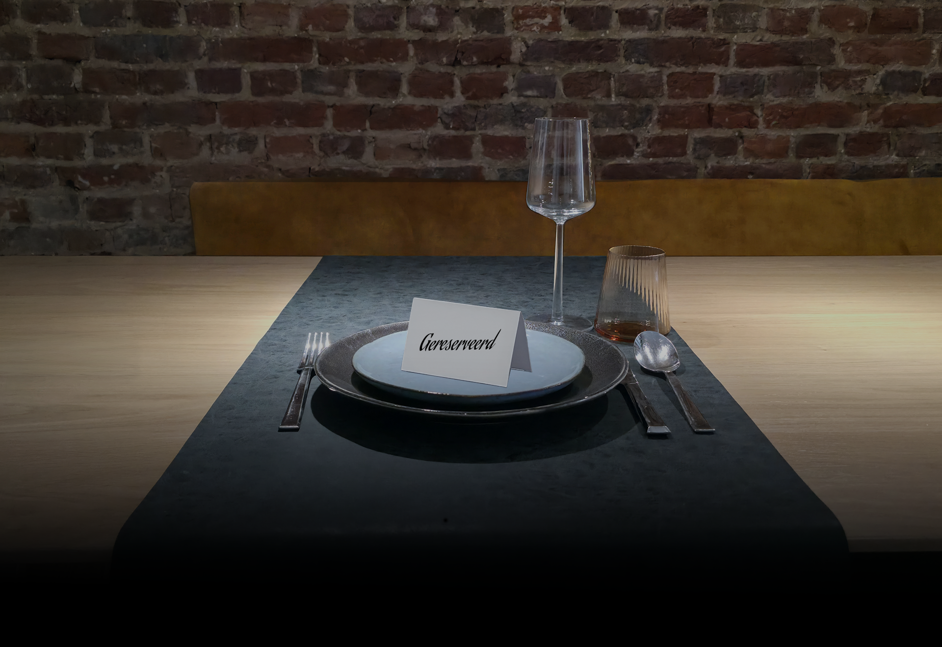 Een foto van de gedekte eettafel bij Michaël en An. Een houten tafel met een zwarte loper waarop borden en glazen staan. Op de achtergrond zie je een okergele zitbank tegen een bakstenen muur. 